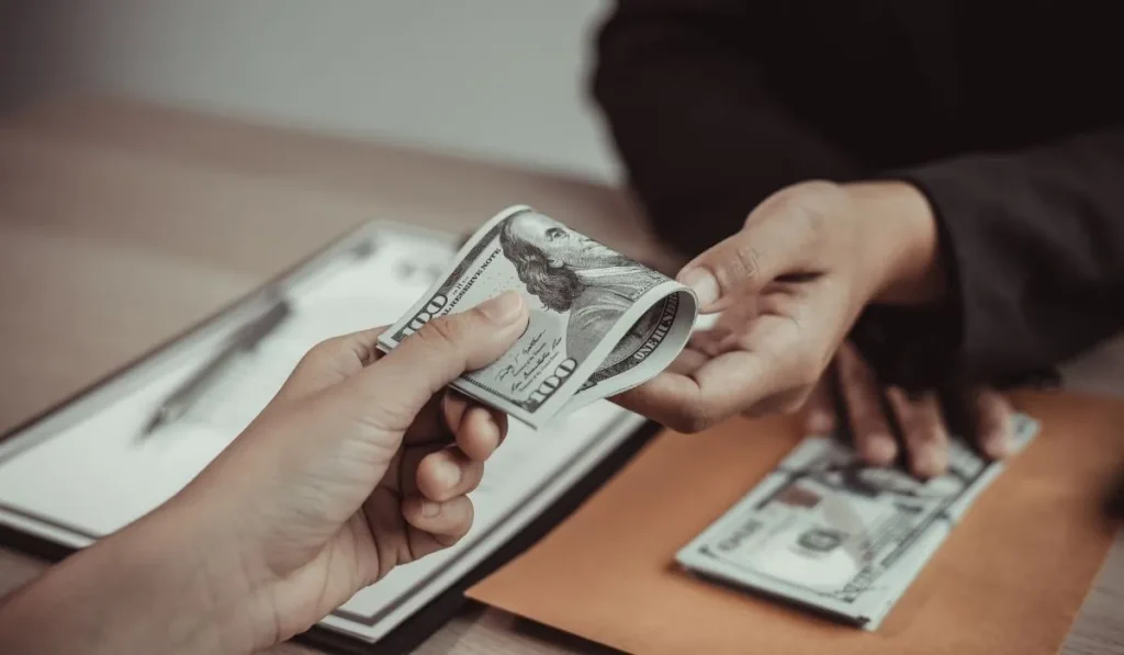 Two people exchanging dollar bills over documents on a table.