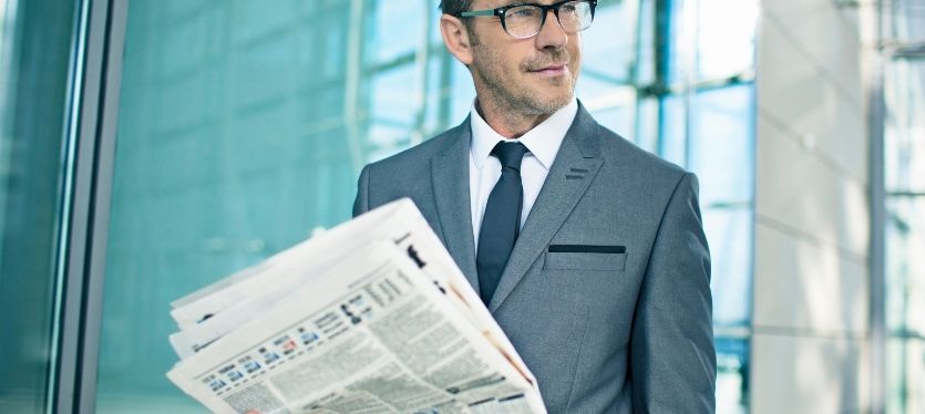 A man wearing a suit and glasses holds a newspaper, suggesting he is informed about current events