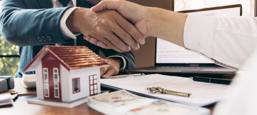 Two people engaged in a handshake above a model house, representing a business deal in real estate