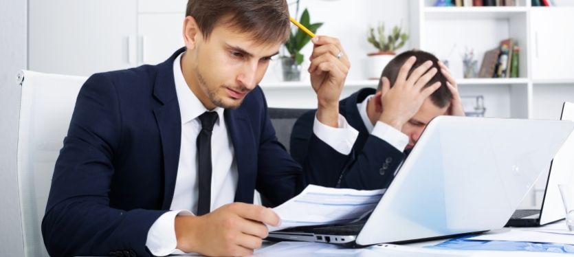 Two suited men collaborating at a desk, each using a laptop in a business environment