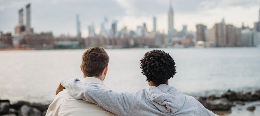 Two men relax on rocks, overlooking a vibrant cityscape, sharing a moment of contemplation