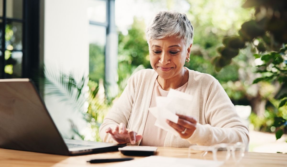 Mujer mayor sentada a una mesa con un portátil y un recibo , mirando sonriente la pantalla al ver que está exonerada de impuesto