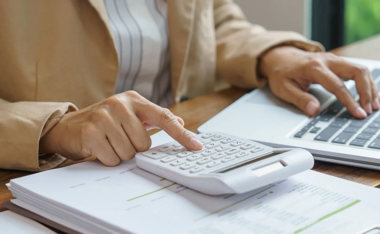 Persona usando una calculadora y laptop en un escritorio, explorando ciudades de Florida para un presupuesto ajustado.