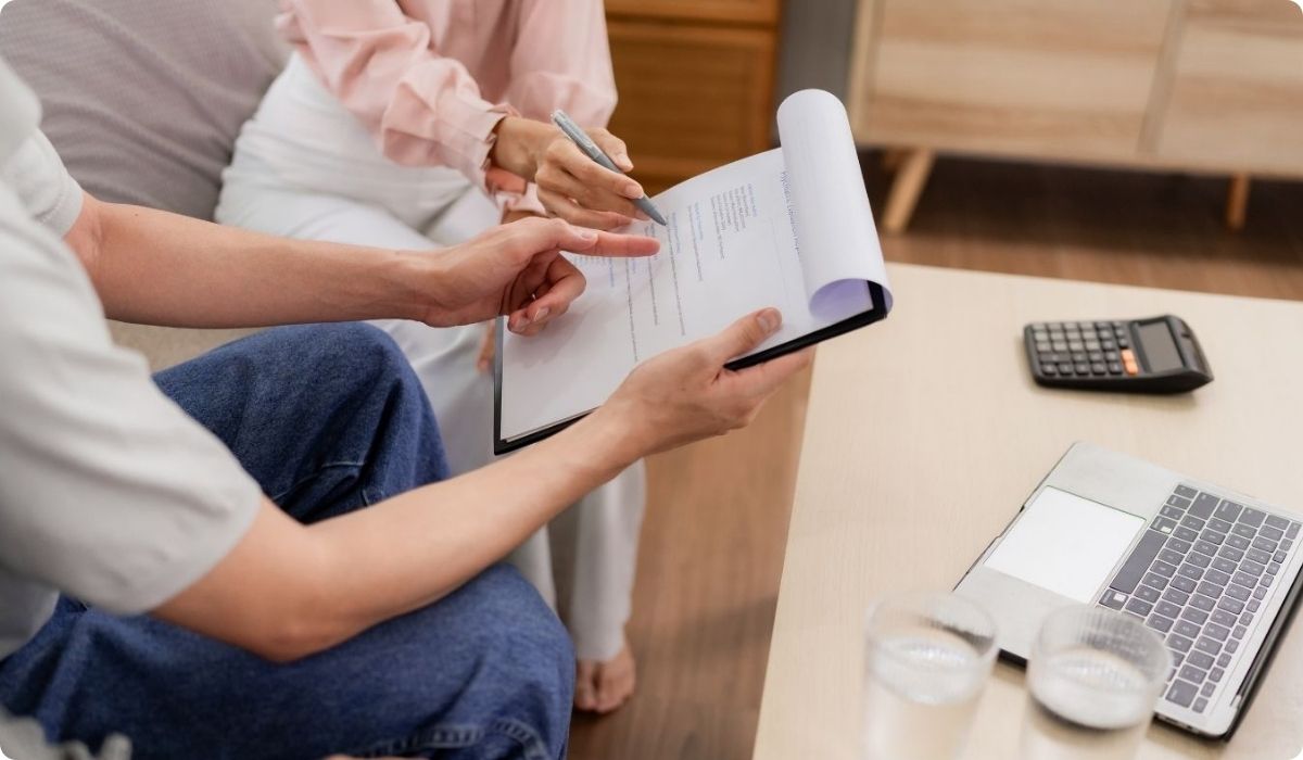 A man and a woman sitting on a sofa, using a laptop and a pen, discussing taxes for Latino investors.