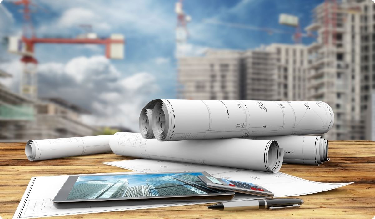 Architectural plans and a tablet on a wooden table, with a construction site in the background, highlighting investment opportunities.