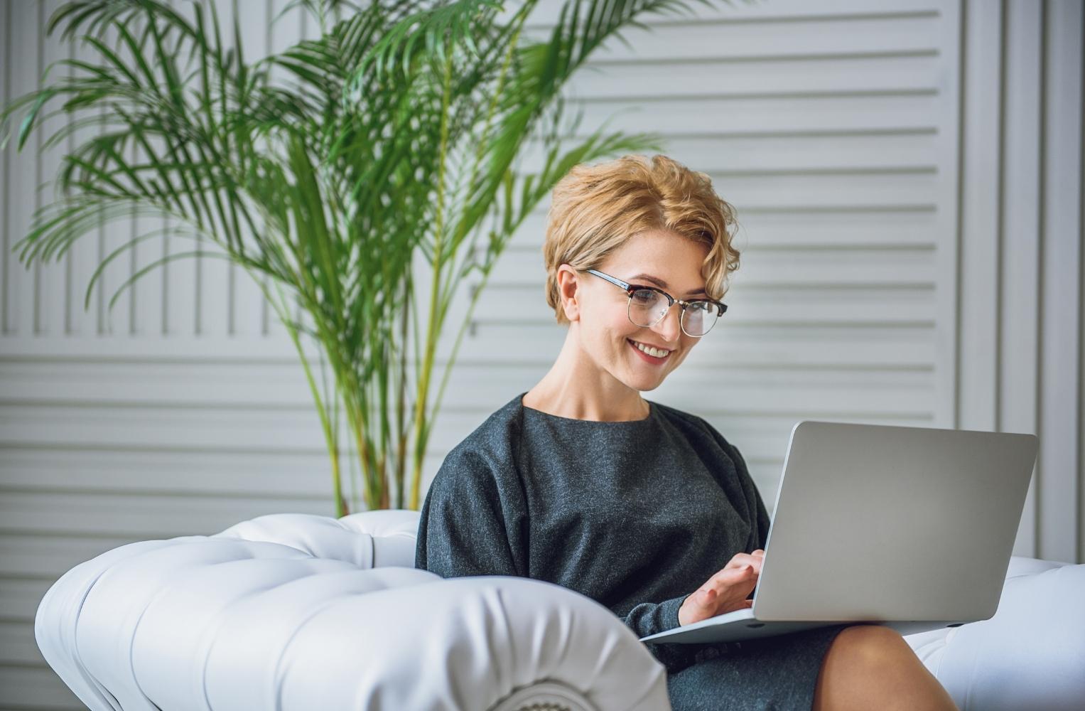  Mujer con gafas sentada en un sofá usando una laptop, enfocada en inversiones en Florida con crédito internacional. 
