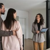 Una pareja frente a una casa, sosteniendo una tableta, mientras un agente inmobiliario les muestra la propiedad.