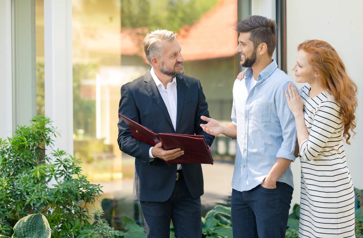 Un agente inmobiliario profesional asesorando a una pareja joven frente a una propiedad.