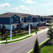 Vista aérea de un complejo de townhomes modernos de dos niveles con fachada azul y blanca, césped verde y calles pavimentadas bajo un cielo despejado.