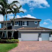 Casa moderna de dos pisos en color gris con techo de tejas terracota, garajes blancos y palmeras bajo un cielo azul brillante.