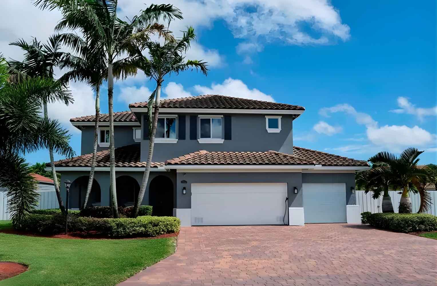 Casa moderna de dos pisos en color gris con techo de tejas terracota, garajes blancos y palmeras bajo un cielo azul brillante.