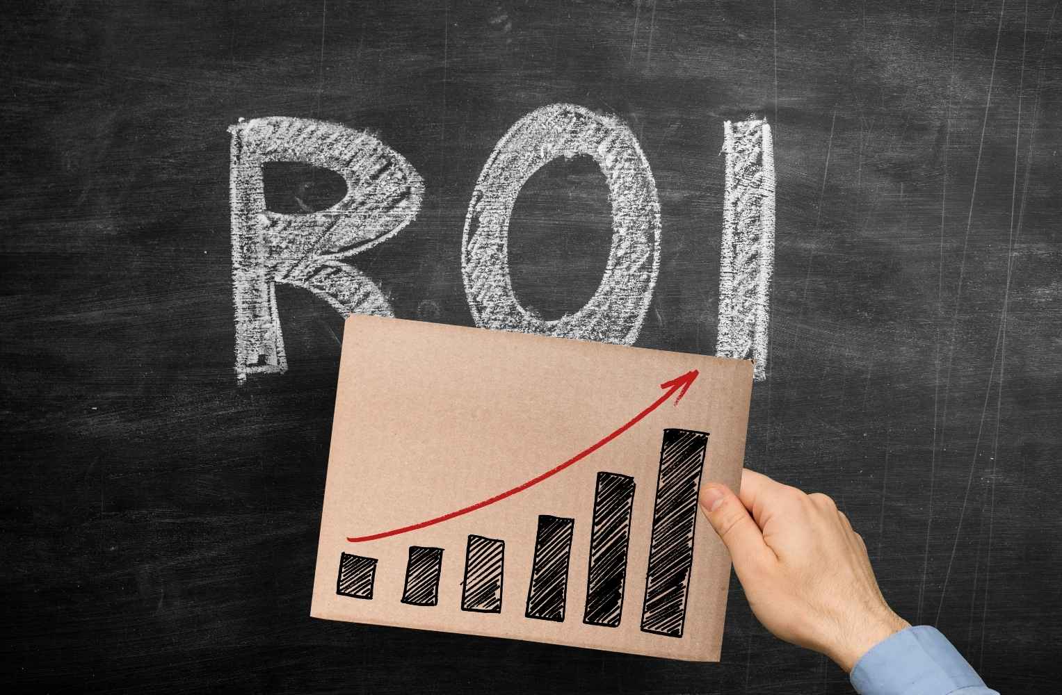 A hand holding a cardboard sign with a rising bar graph and red arrow in front of a chalkboard with "ROI" written in white chalk.