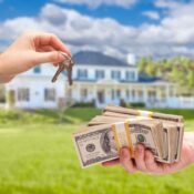 Una mano entregando un juego de llaves mientras otra sostiene varios fajos de billetes de cien dólares, con una casa blanca desenfocada al fondo.