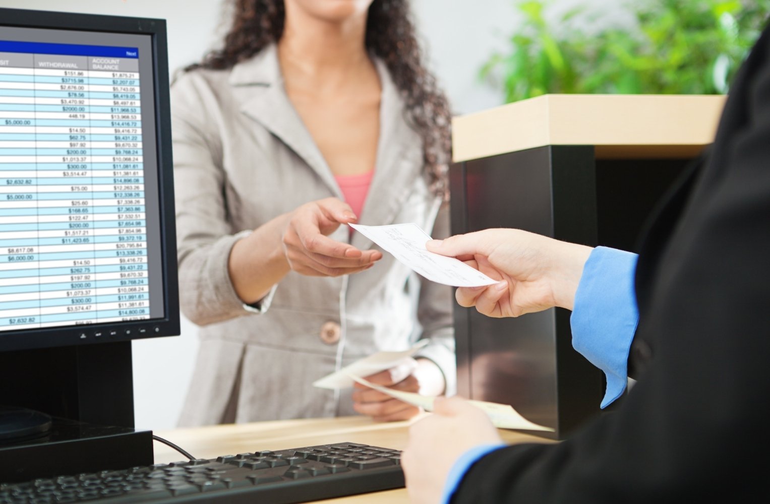 Mujer entregando un cheque a otra en una oficina bancaria con una pantalla de computadora que muestra tablas financieras de fondo.