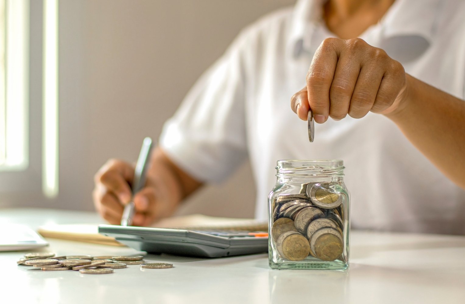 Person saving coins in a glass jar next to a calculator.