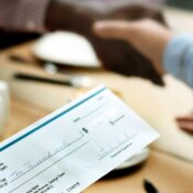 Primer plano de un cheque de depósito de diez mil dólares sobre una mesa con personas dándose la mano al fondo.