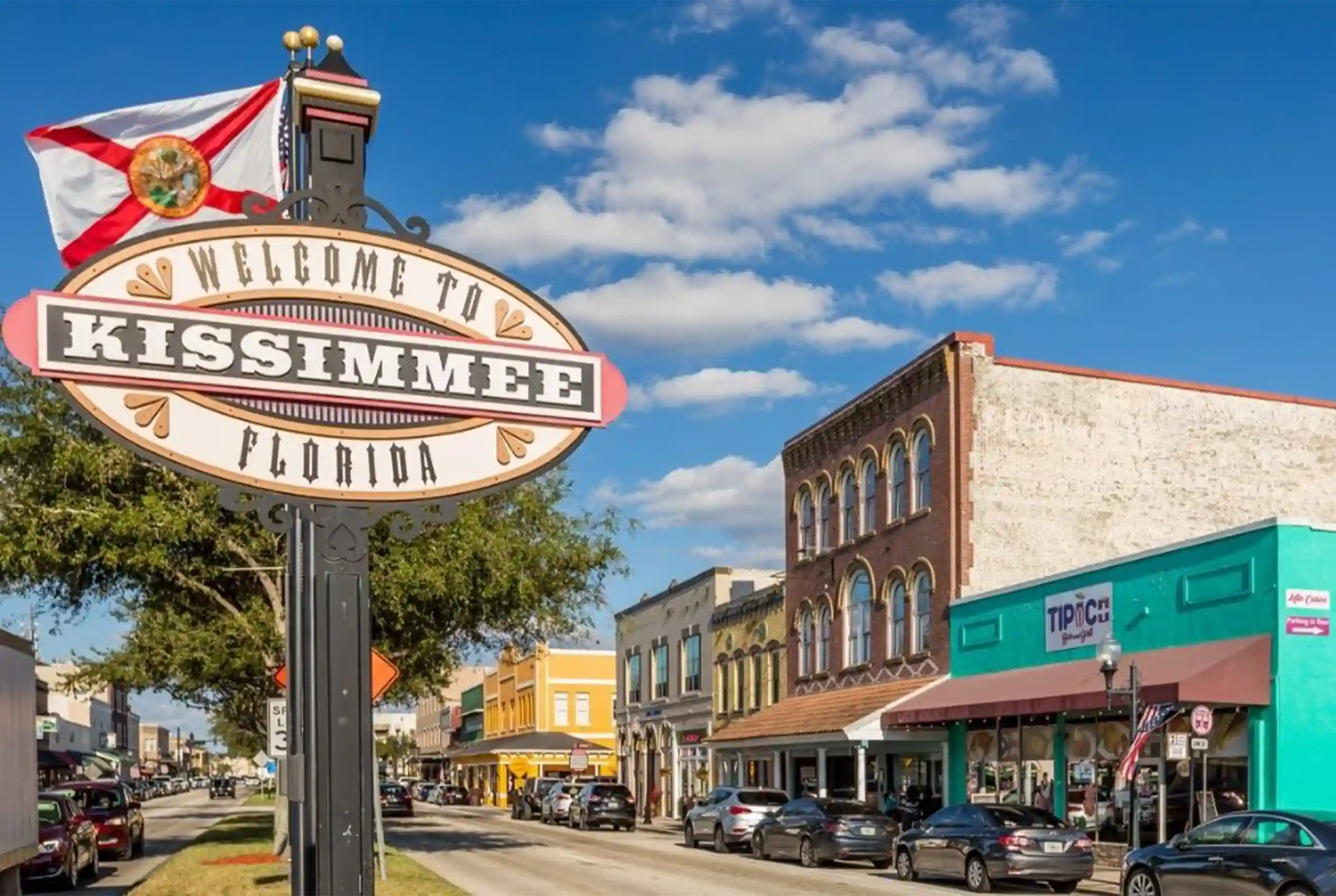 Cartel de bienvenida a Kissimmee, Florida, con la bandera del estado y el downtown histórico al fondo.