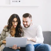 Pareja latinoamericana emocionada buscando propiedades en Florida desde casa, con laptop y letrero de hogar al fondo
